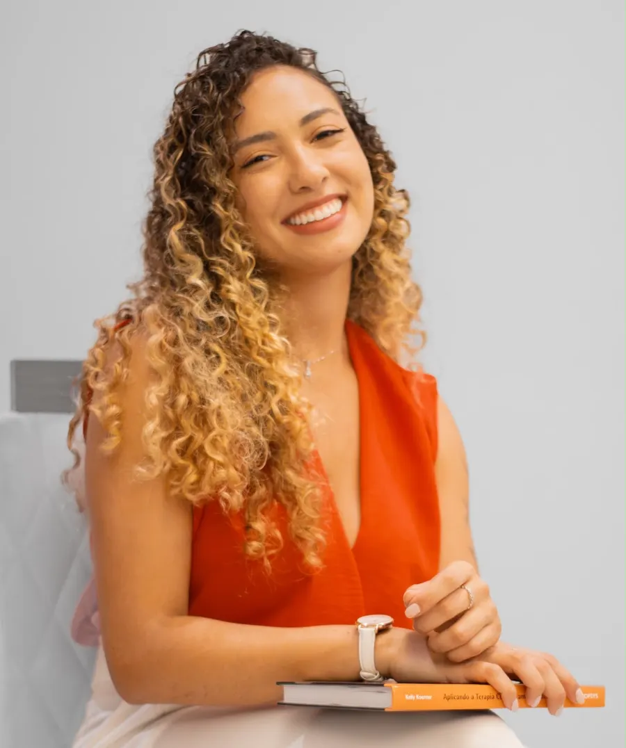 Psicóloga Isabella Lacerda sorrindo, sentada, com um livro de Terapia Cognitivo-Comportamental no colo